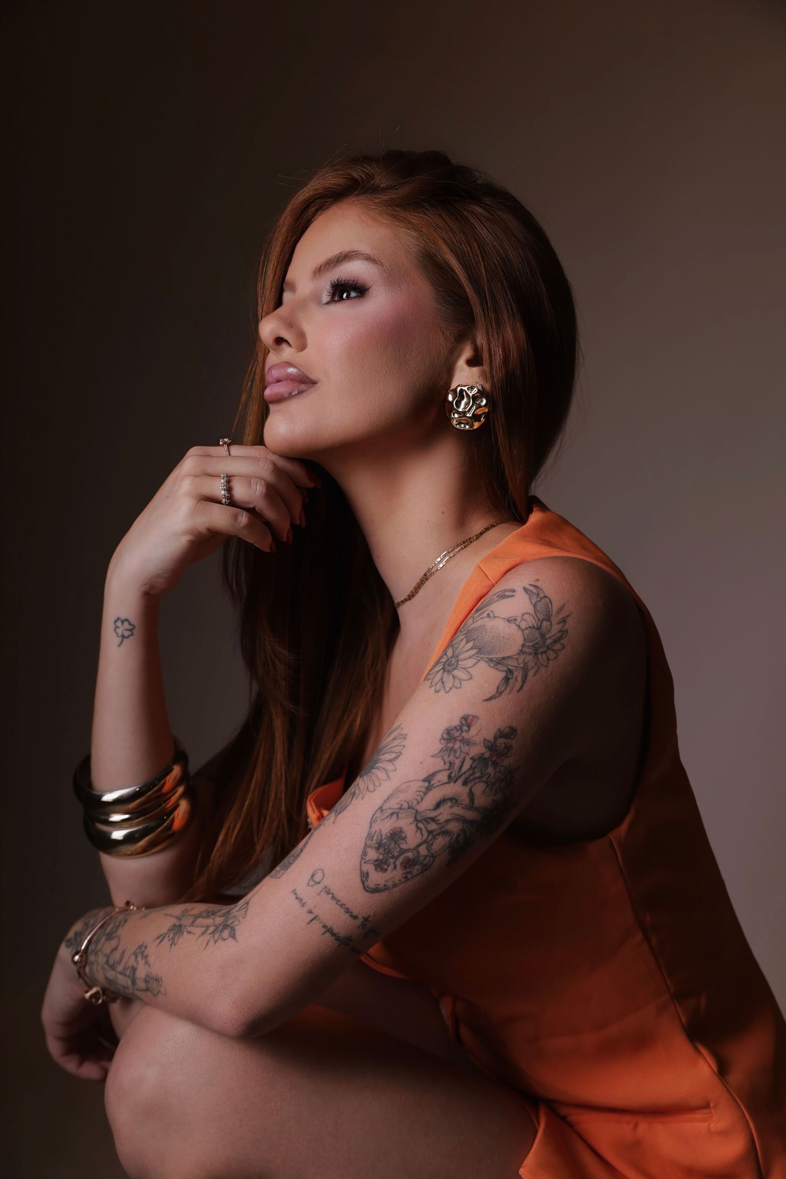 Dra. Mariah Pires de perfil, com roupa laranja e tatuagens no braço, em fundo escuro.